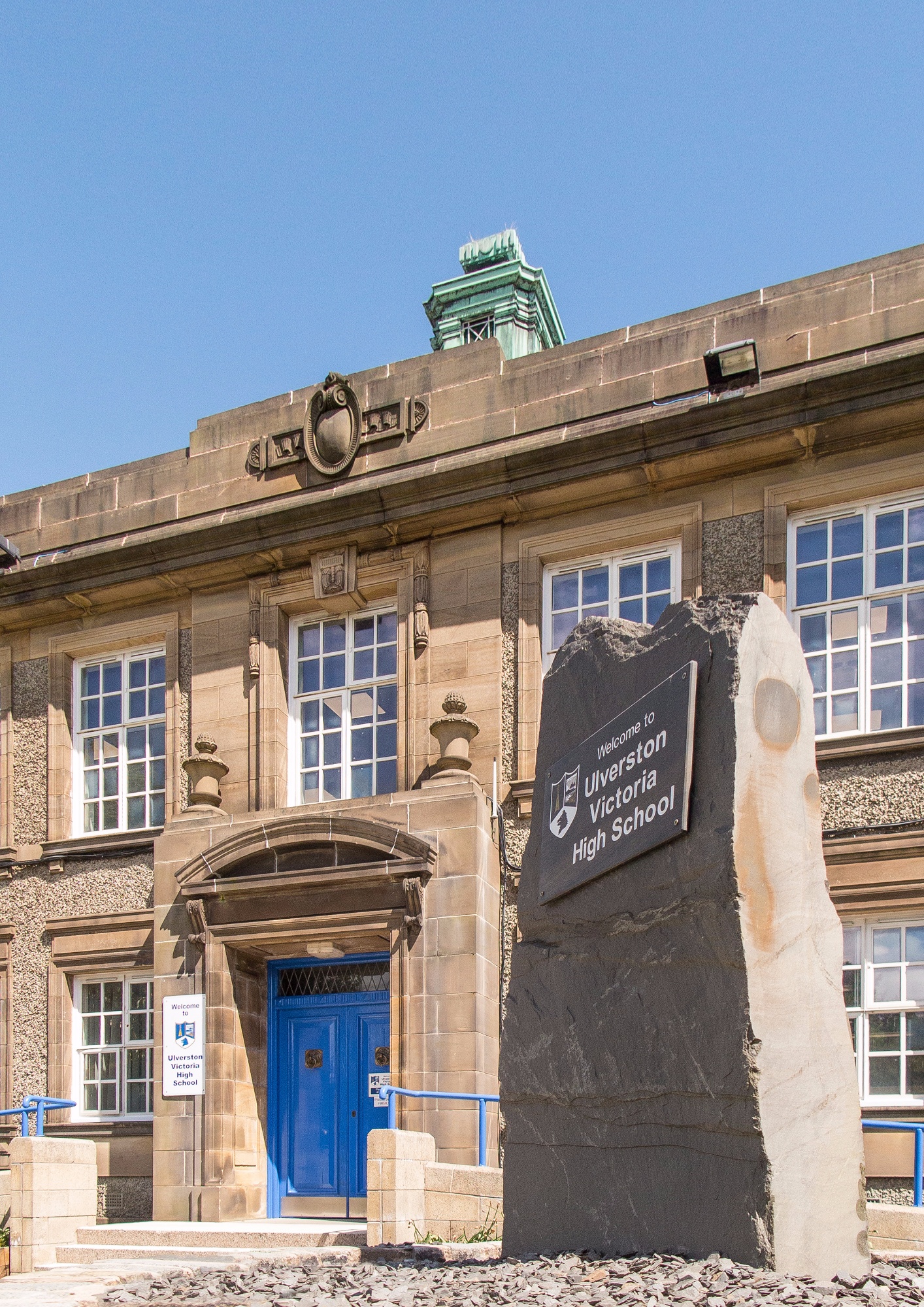Stone school front with slate column in front with the school name and logo.